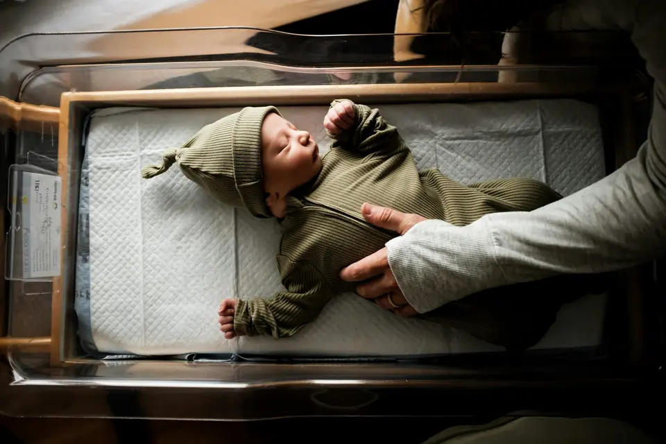 A sleeping newborn in a knit hat and sleeper lying in a hospital bassinet, with a parent's hand resting on him