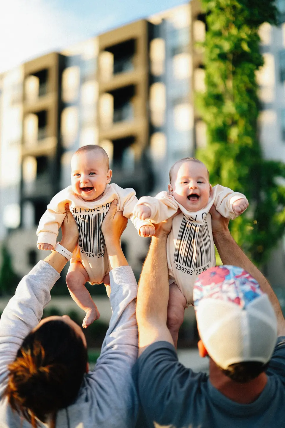 Two parents joyfully lifting their laughing, wide-awake twin babies in golden-hour light