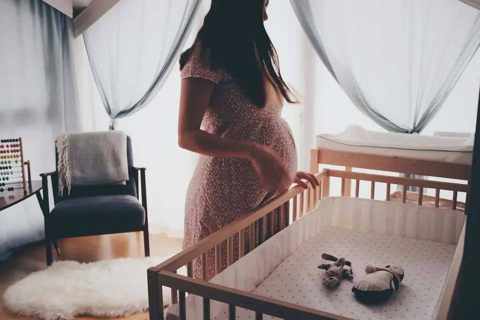 A pregnant woman standing at a crib in a sunlit nursery, back to camera