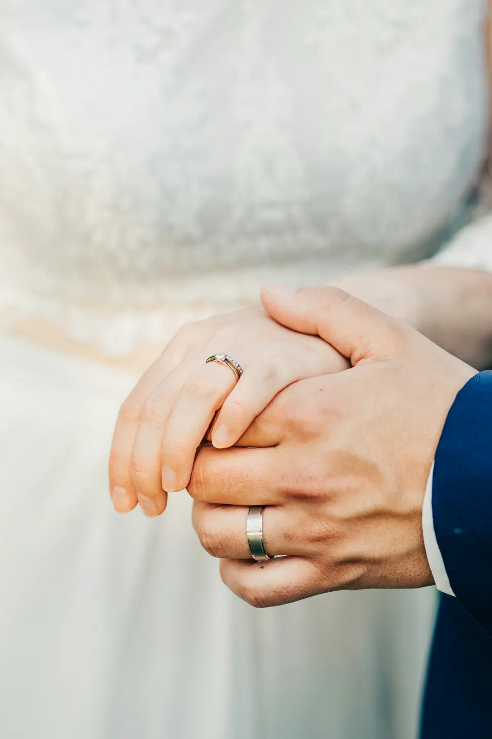 A bride and groom's clasped hands, his plain wide wedding band in focus