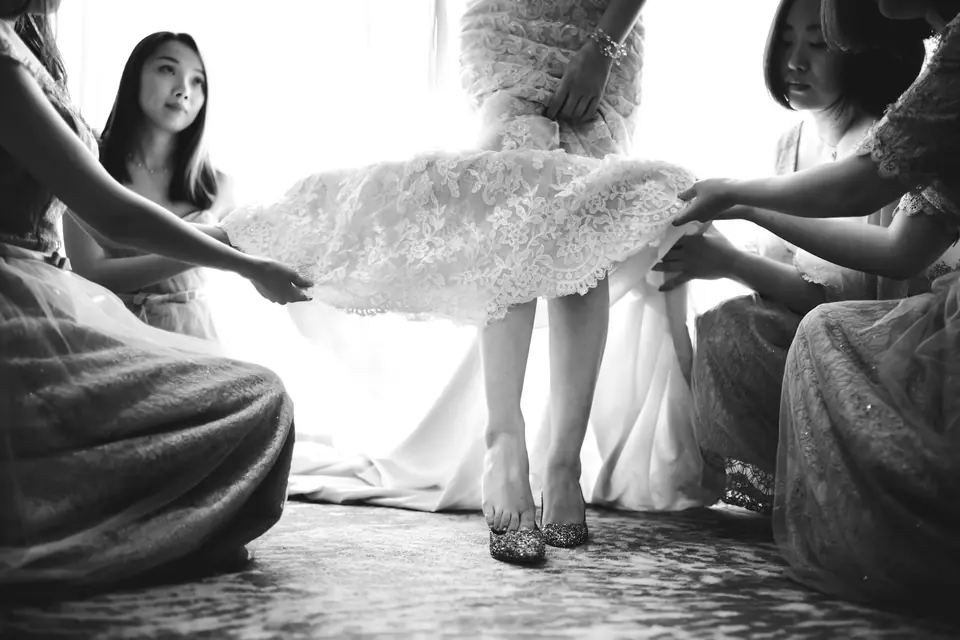 Four bridesmaids holding up the lace hem of a wedding dress while the bride steps into glitter heels, in black and white