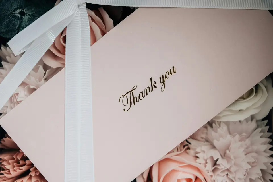 A pale pink thank-you card tied with a white grosgrain ribbon over soft pink roses
