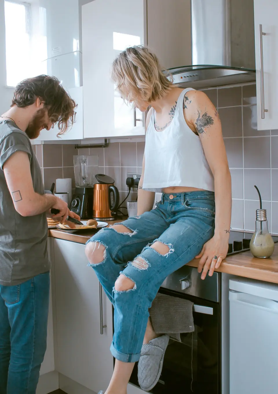 A couple in a kitchen, one slicing fruit at the counter, the other sitting on the counter beside them