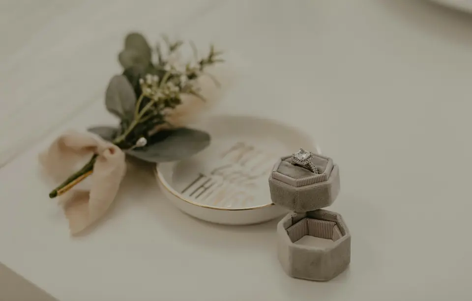 An engagement ring with a square center stone in an open grey velvet ring box, beside a gold-rimmed ring dish and a boutonniere